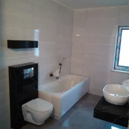 Nowoczesna łazienka z białymi płytkami imitującymi marmur, wisząca toaleta z czarnym marmurowym stelażem, okrągła umywalka nablatowa na czarnym blacie i biała wanna.