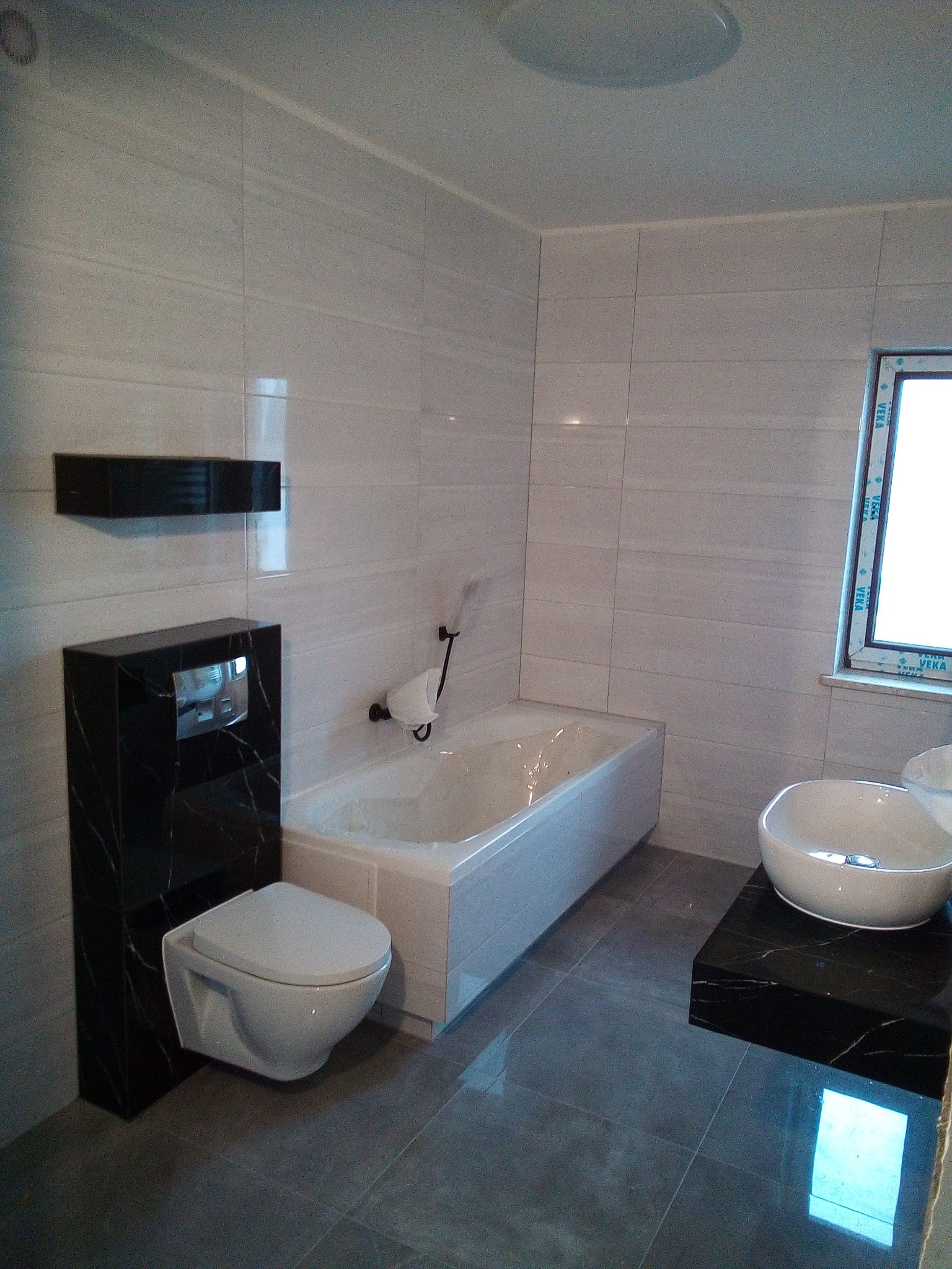 Nowoczesna łazienka z białymi płytkami imitującymi marmur, wisząca toaleta z czarnym marmurowym stelażem, okrągła umywalka nablatowa na czarnym blacie i biała wanna.