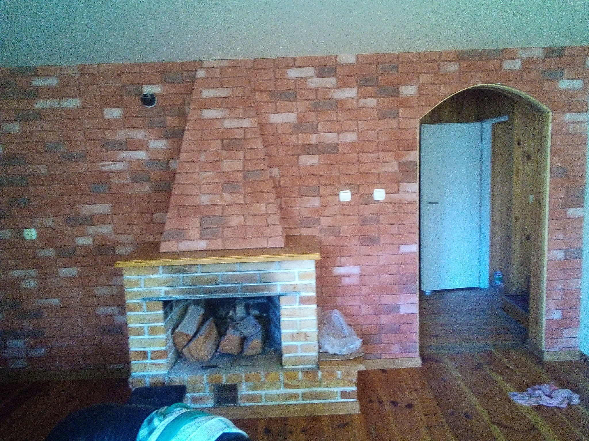 Ceglana ściana z kominkiem i łukowym przejściem do innego pomieszczenia, drewniana podłoga, fragment mebla w rogu