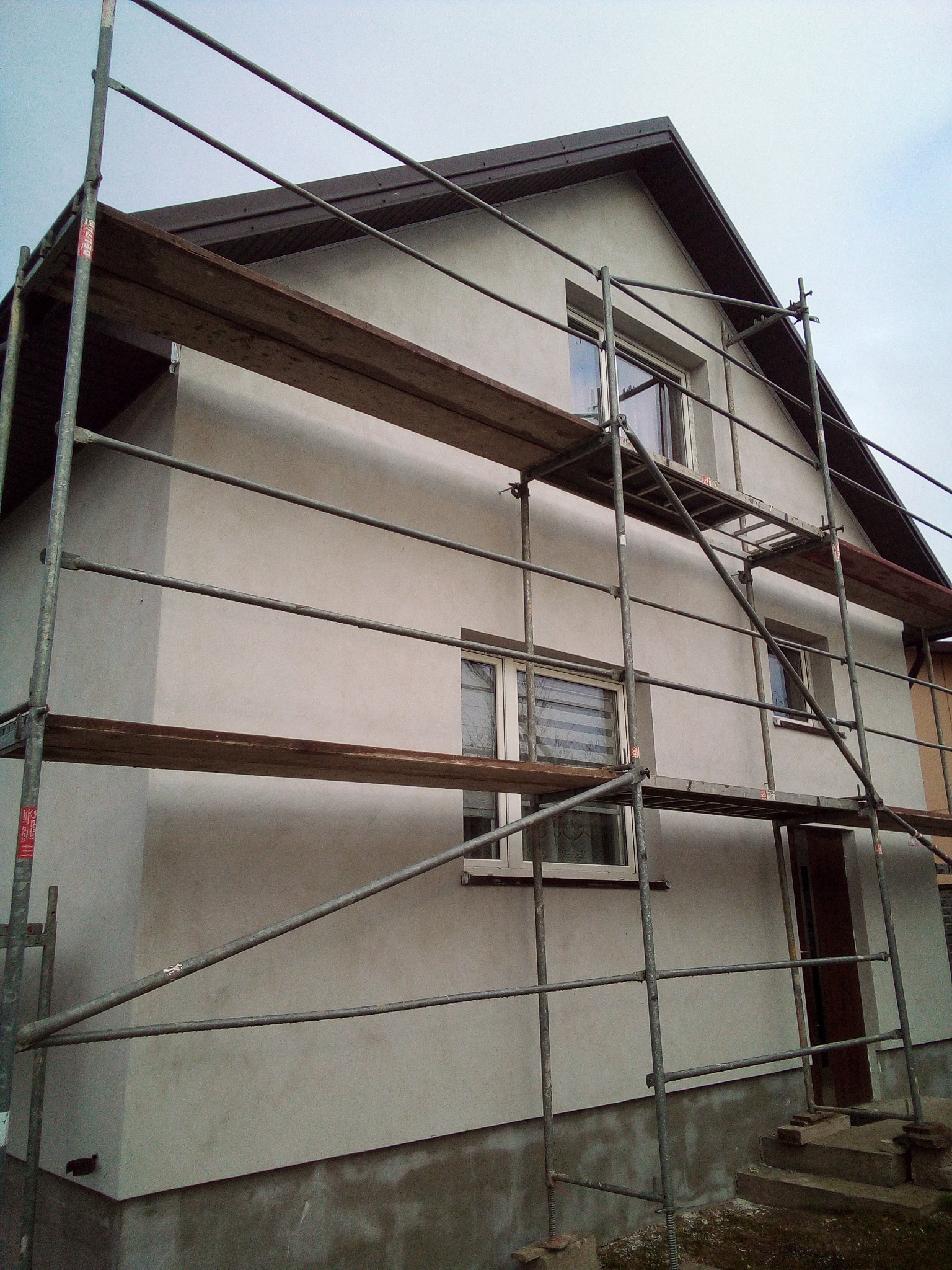 Dom jednorodzinny w trakcie prac elewacyjnych, obłożony rusztowaniem, świeżo otynkowany na szaro, z widocznymi oknami i wejściem.