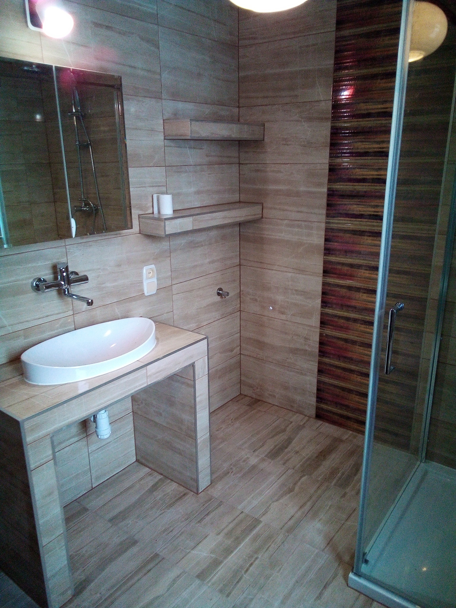 Nowoczesna łazienka w Małogoszczu z kabiną prysznicową, umywalką nablatową na postumencie z płytek w odcieniach beżu i brązu oraz półkami ściennymi.