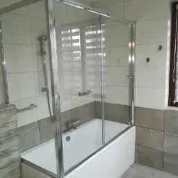 Biała wanna z zamontowaną szklaną kabiną prysznicową z chromowanymi profilami, na tle jasnych płytek w łazience.