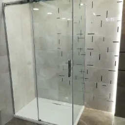 Kabina prysznicowa z przesuwnymi drzwiami i chromowanymi profilami, osadzona na białym brodziku, na tle płytek z geometrycznym wzorem.