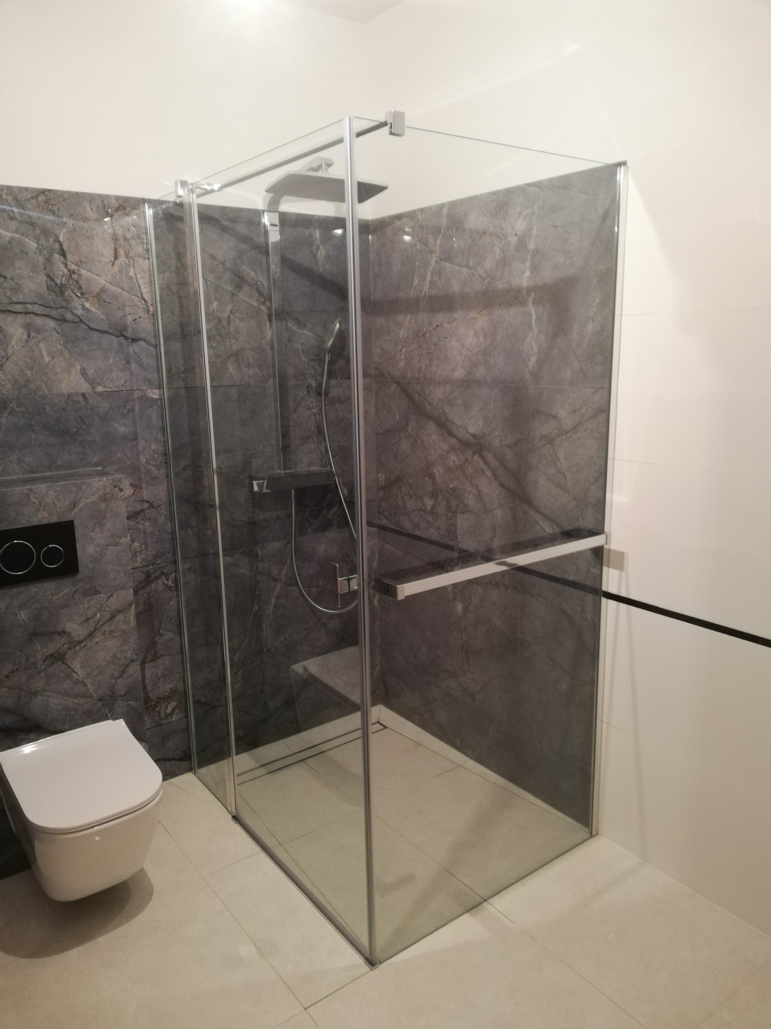 Nowoczesna kabina prysznicowa z deszczownicą i szklaną półką, obok podwieszana toaleta na tle marmurowych płytek w łazience w Mrągowie.