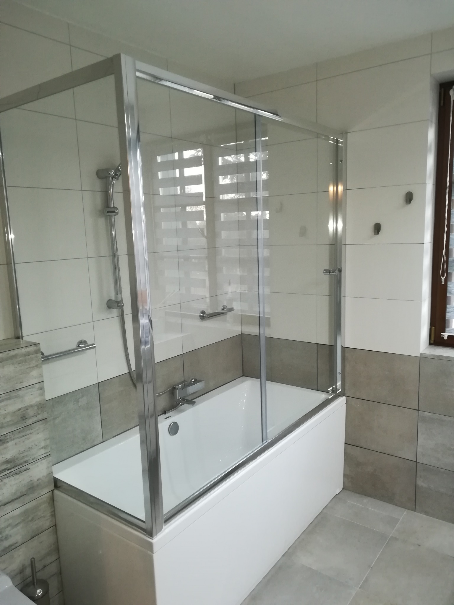 Biała wanna z zamontowaną szklaną kabiną prysznicową z chromowanymi profilami, na tle jasnych płytek w łazience.