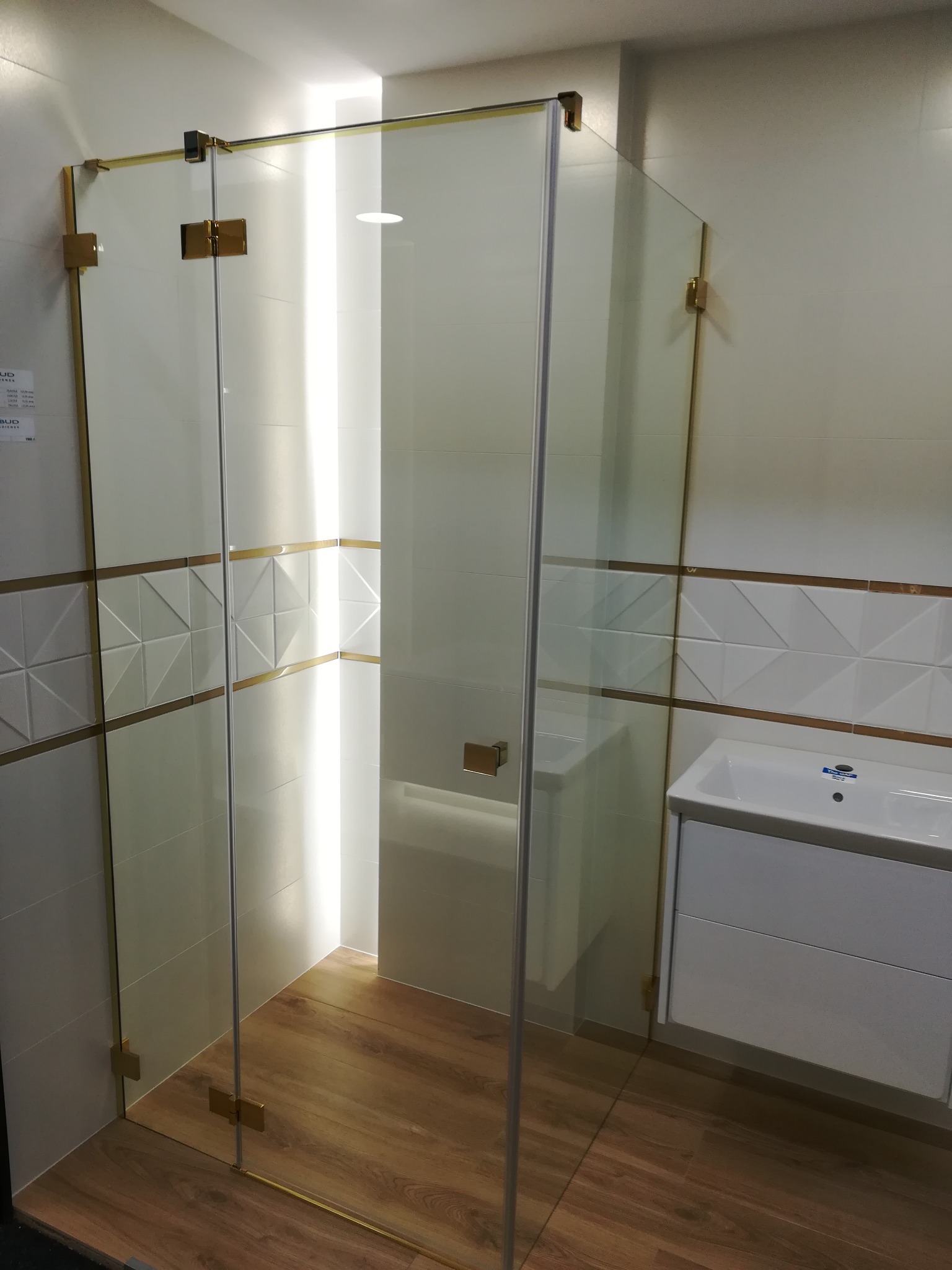 Kabina prysznicowa ze złotymi okuciami w łazience z drewnopodobną podłogą i białymi płytkami z geometrycznym wzorem.