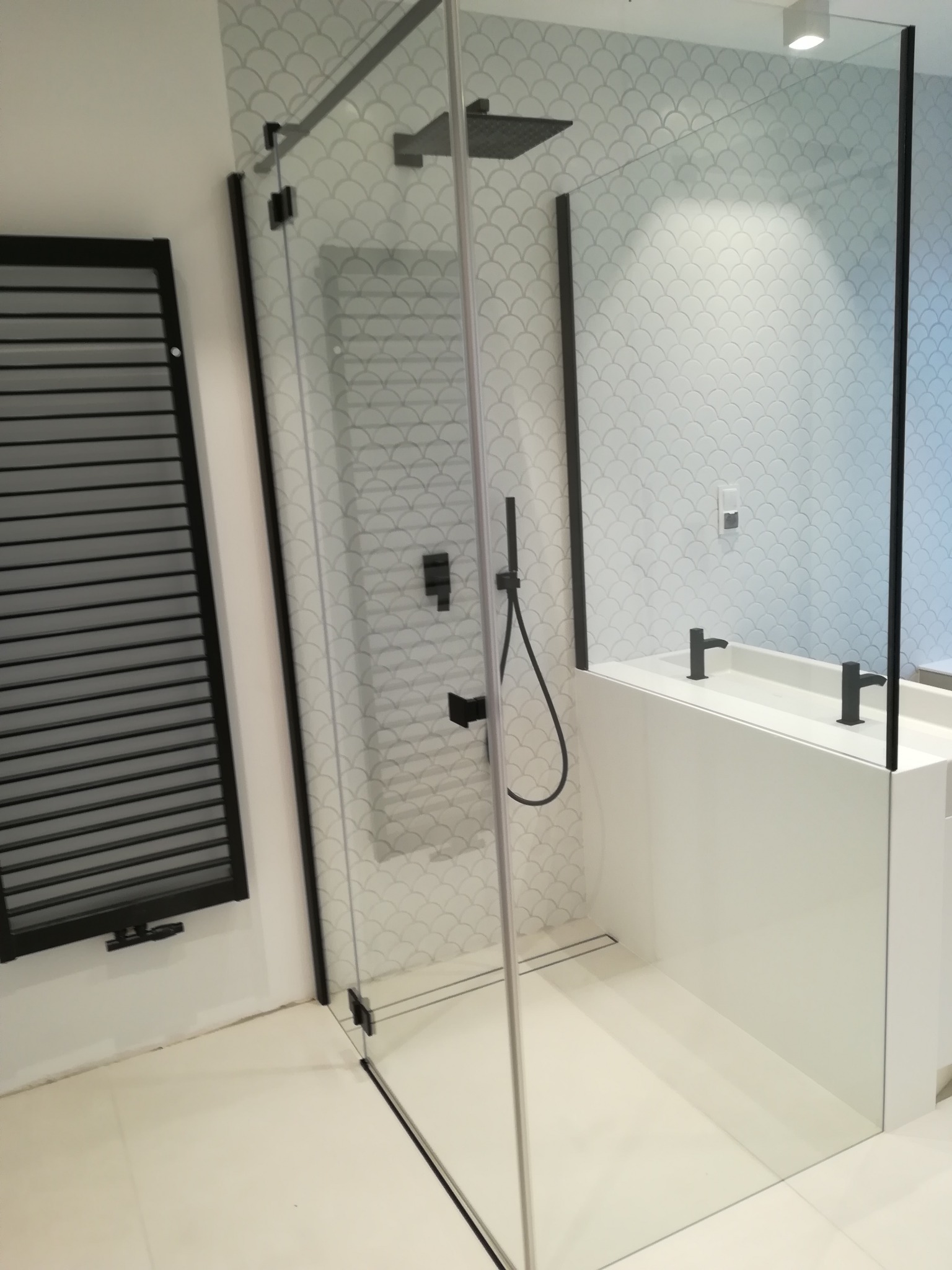 Nowoczesna łazienka z kabiną prysznicową typu walk-in z czarnymi profilami, grzejnikiem drabinkowym i umywalką nablatową z dwoma czarnymi bateriami.