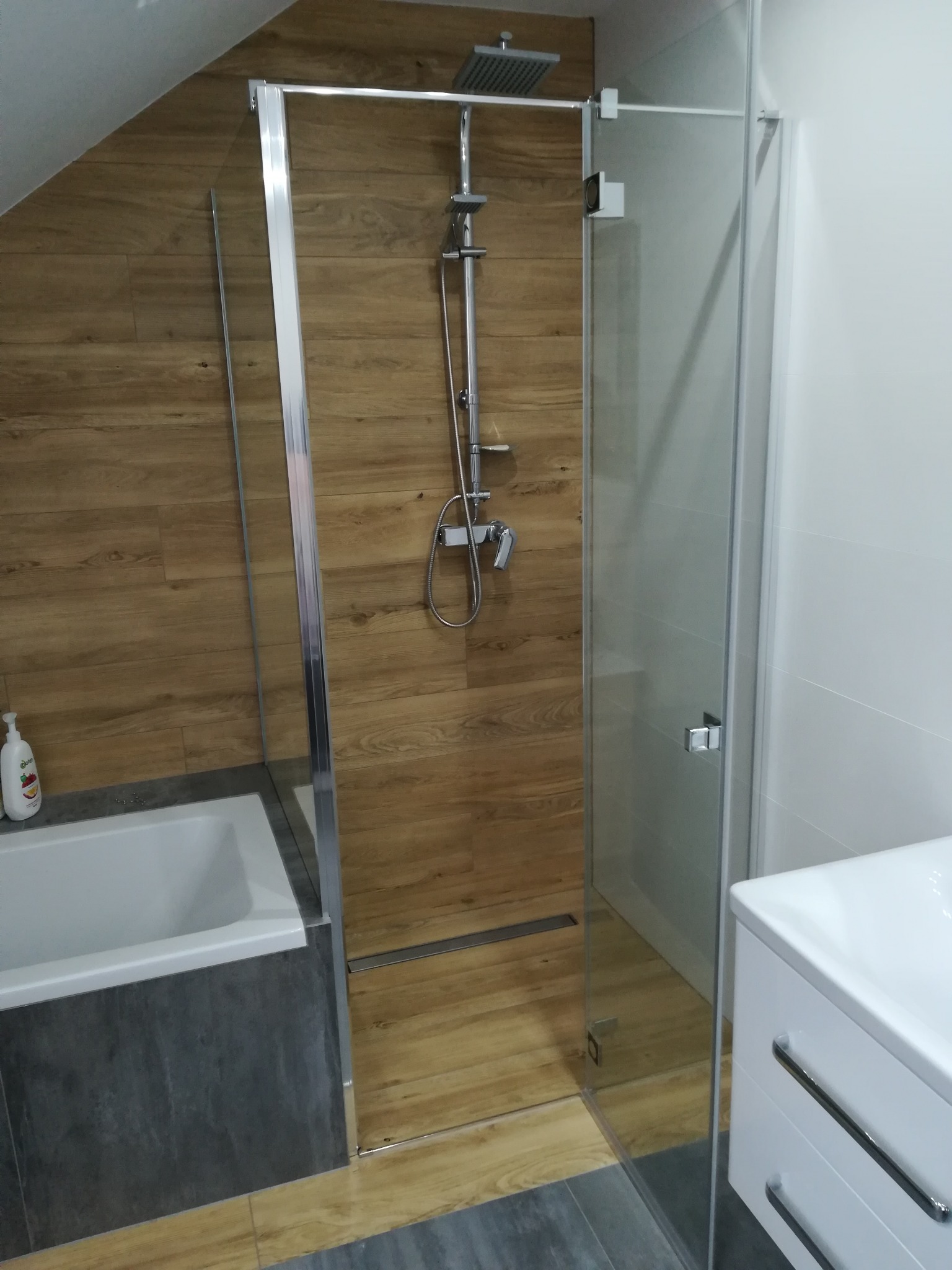 Kabina prysznicowa z deszczownicą, drewnopodobne płytki na ścianie, biała umywalka z szafką, szare płytki podłogowe, widok na odpływ liniowy.