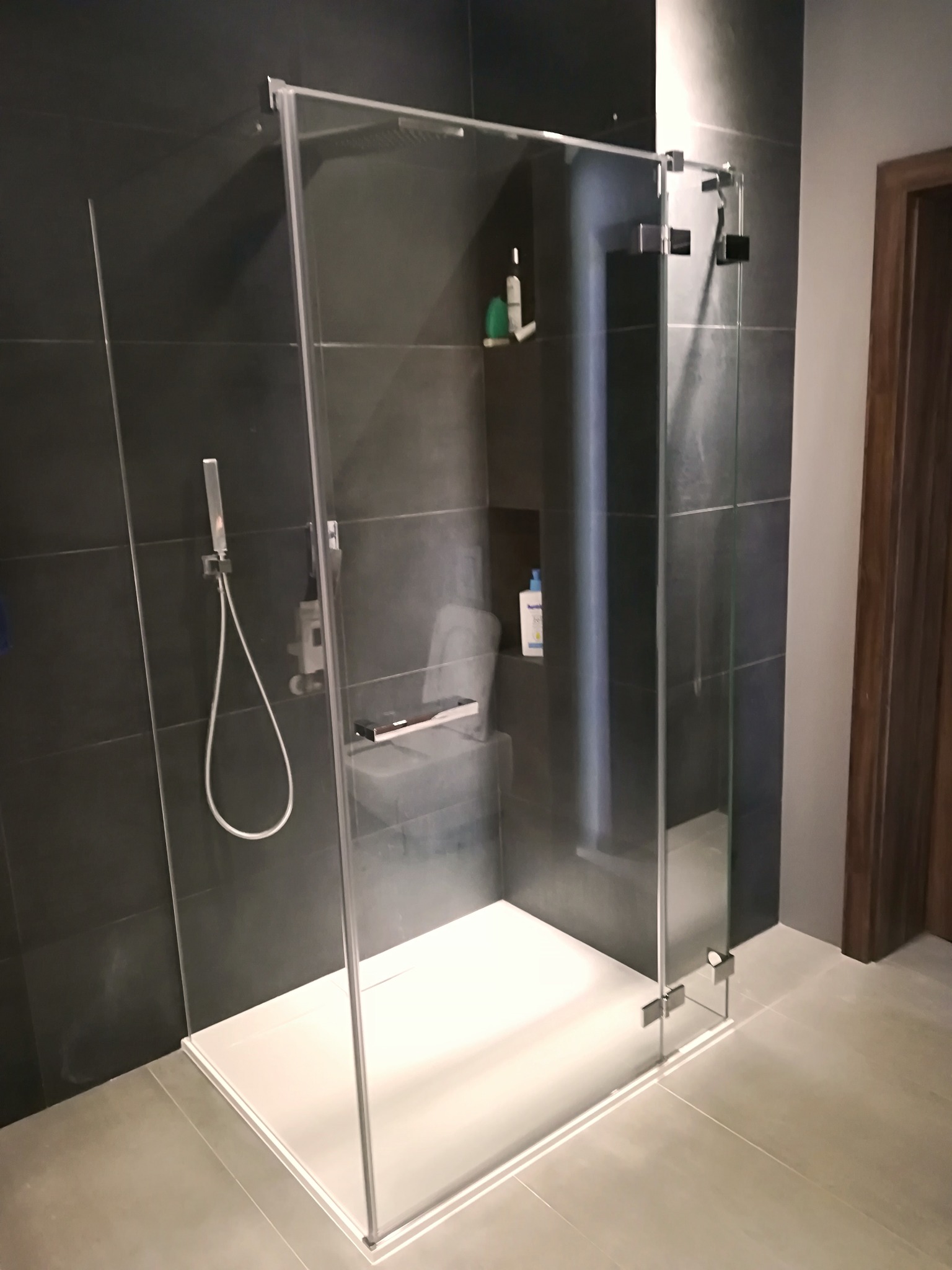 Nowoczesna kabina prysznicowa z transparentnego szkła, czarne płytki na ścianach, biały brodzik, bateria prysznicowa z deszczownicą i półka z kosmetykami w tle.