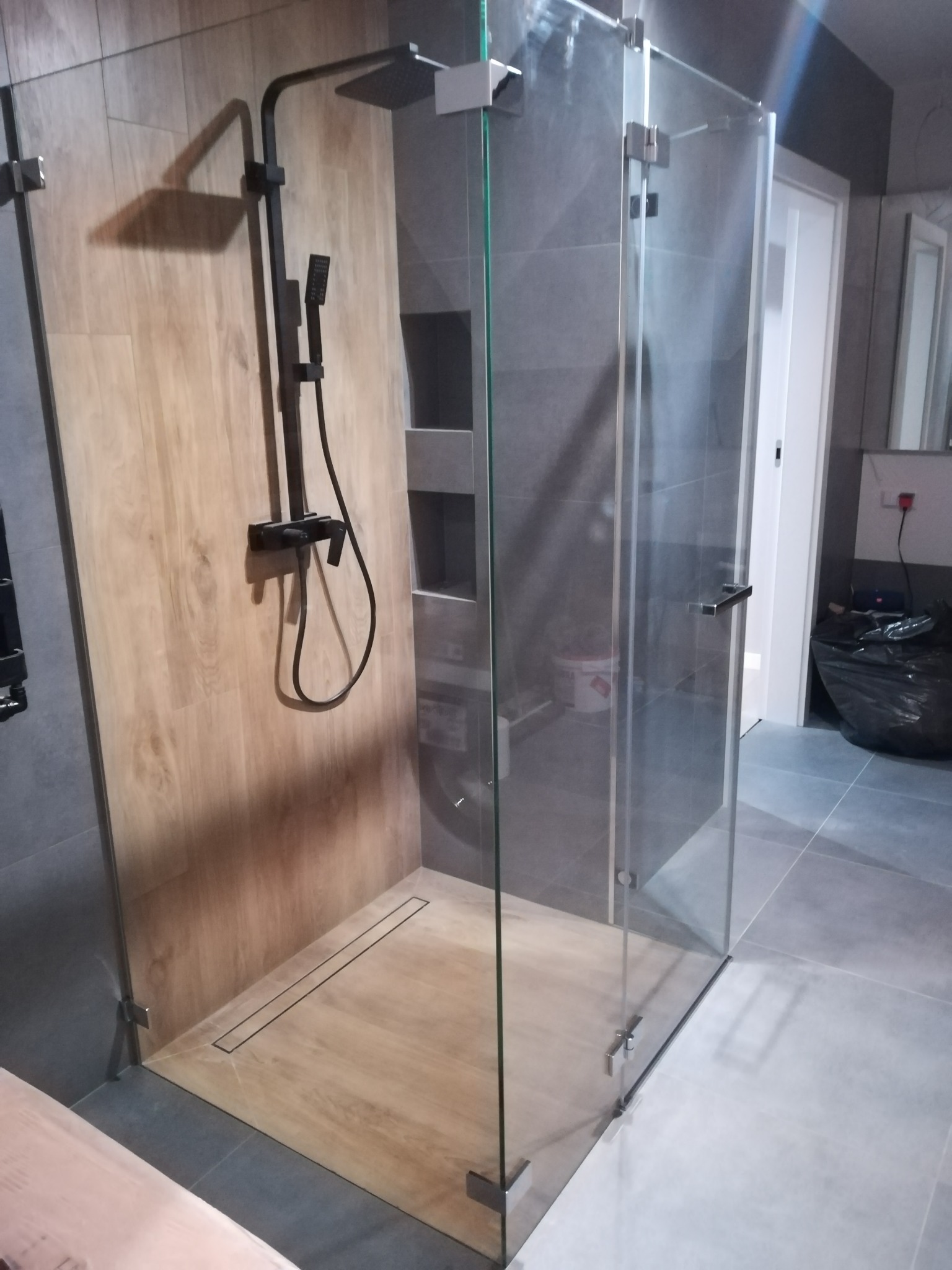 Nowoczesna kabina prysznicowa typu walk-in z czarną armaturą, wykończona płytkami imitującymi drewno i szarymi płytkami wielkoformatowymi, z widocznym odpływem liniowym.