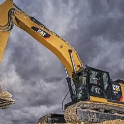 Żółta koparka gąsienicowa marki CAT na tle burzowego nieba, operator w kabinie, łyżka uniesiona w górę, widoczne ślady ziemi na łyżce.