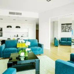 Jasne, przestronne wnętrze apartamentu z turkusową sofą i fotelem, czarnym stolikiem kawowym, białymi meblami kuchennymi w tle i drewnianą podłogą.