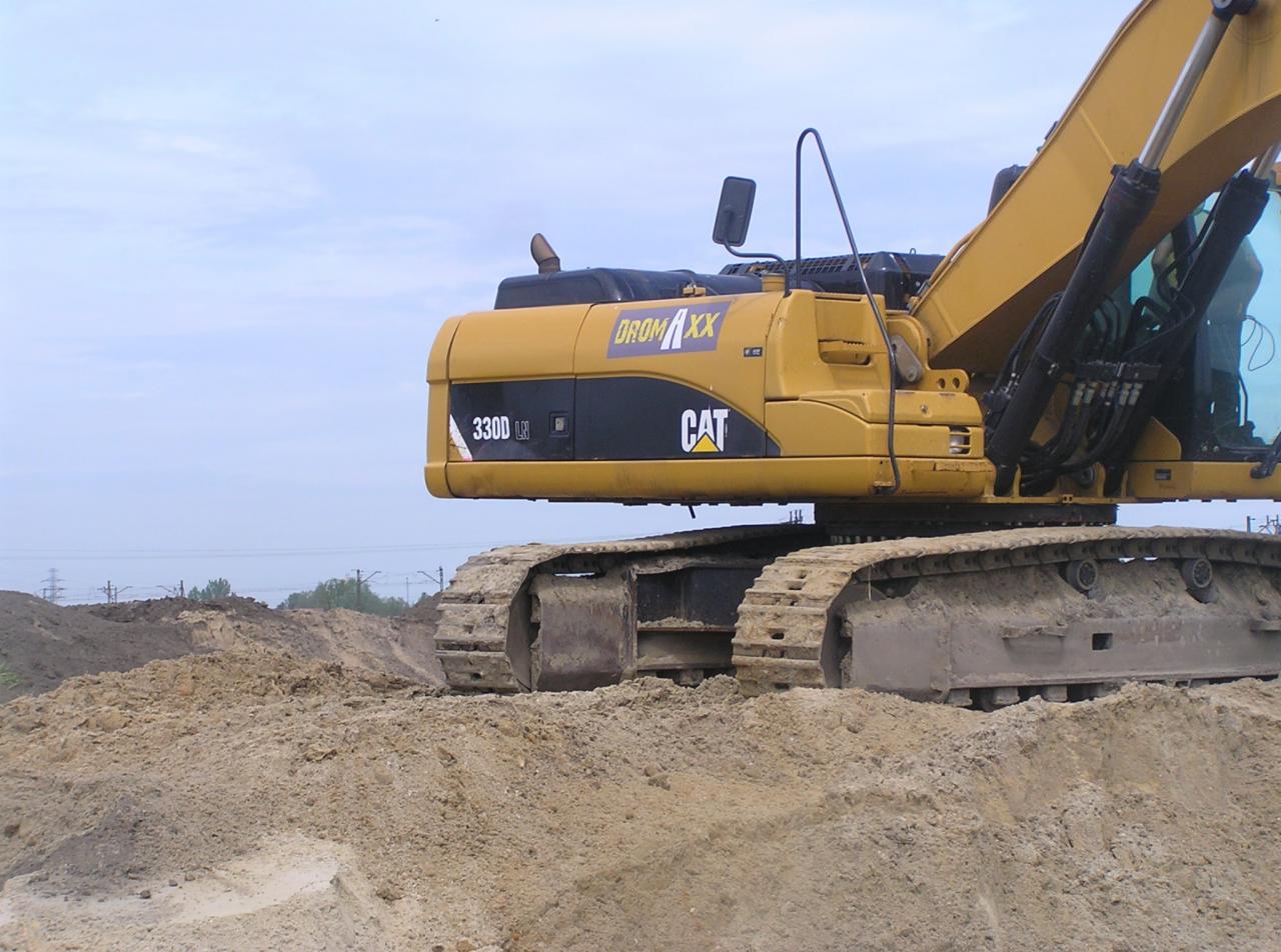 Żółta koparka gąsienicowa marki CAT 330D LX na placu budowy, widoczna na tle sterty ziemi i słupów wysokiego napięcia w oddali.