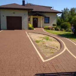 Ułożona brązowa kostka betonowa tworząca podjazd i ścieżkę do domu z beżową elewacją i brązowym dachem, z wydzielonym obszarem na klomb w kształcie półksiężyca.