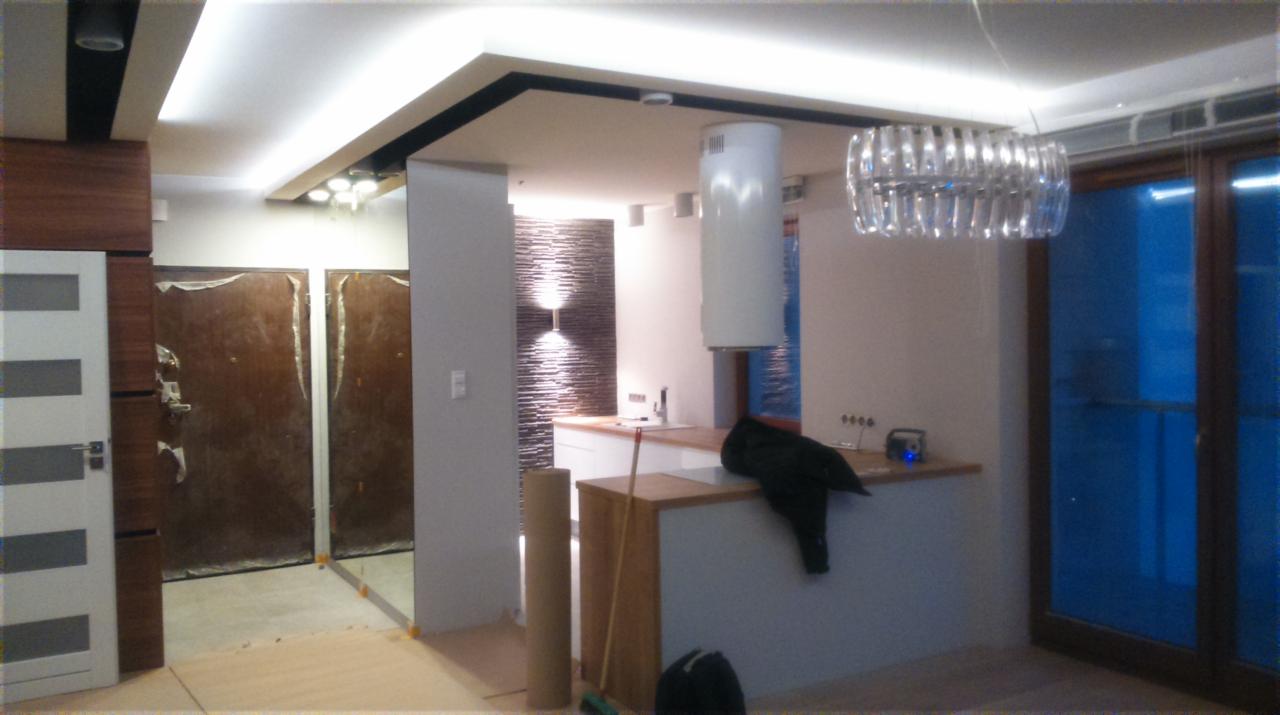 Wnętrze po remoncie: kuchnia z wyspą i wykończoną ścianą z dekoracyjnej cegły. Widoczne zabezpieczone drzwi i nowoczesne oświetlenie.