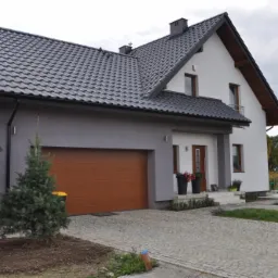 Dwukondygnacyjny dom z szarą elewacją, brązową bramą garażową i czarną dachówką, widoczny z perspektywy podjazdu wyłożonego kostką brukową, z małą choinką w pierwszym planie.