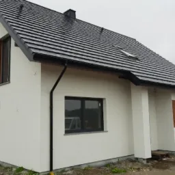 Nowoczesny, jednorodzinny dom z białym tynkiem, ciemną dachówką i drewnianymi panelami dekoracyjnymi na elewacji, widoczny z zewnątrz w pochmurny dzień.