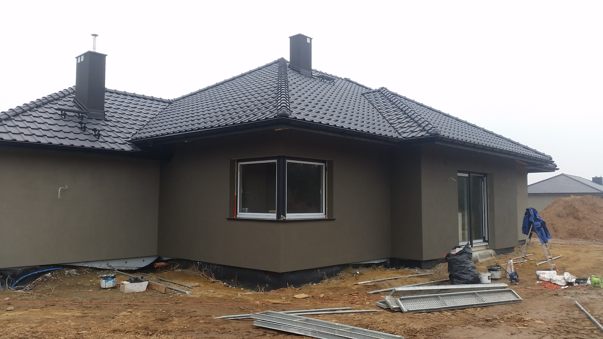 Nowoczesny dom z szarą elewacją i grafitową dachówką, widoczne rusztowania i materiały budowlane wokół, sugerujące trwające prace wykończeniowe.