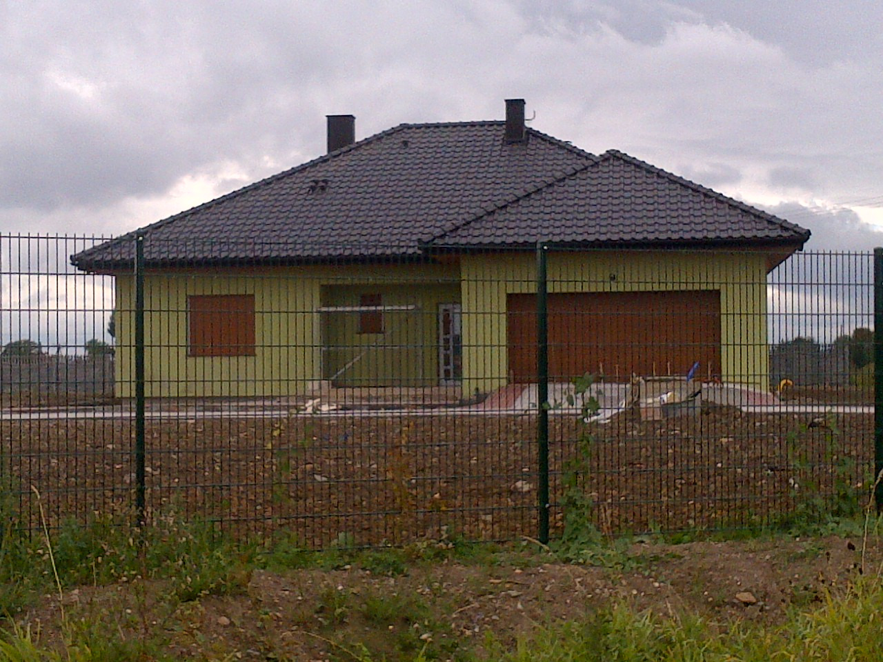 Żółty dom jednorodzinny z ciemną dachówką, widoczny przez metalowe ogrodzenie, w trakcie wykończenia terenu wokół budynku.