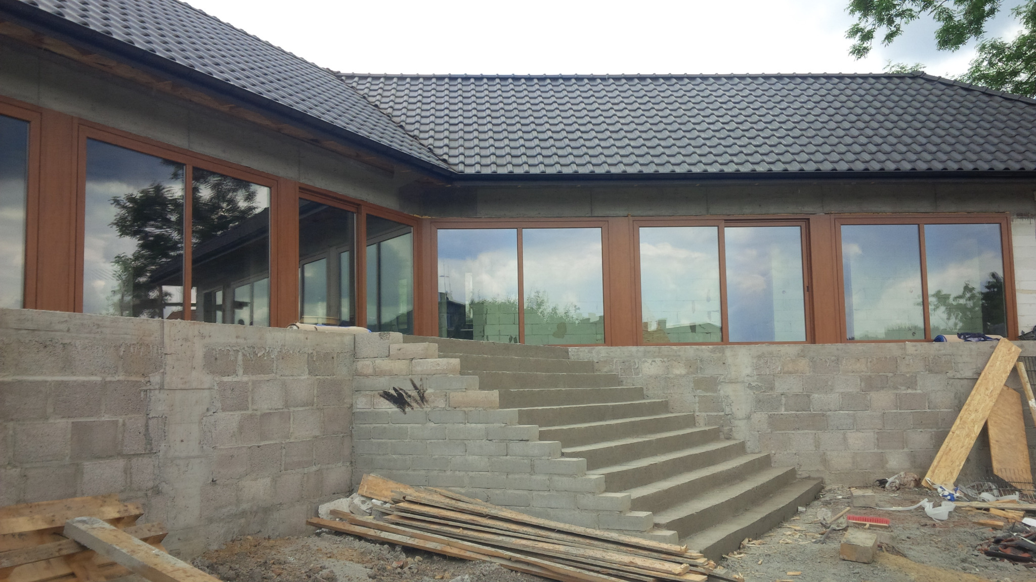 Budynek w trakcie rozbudowy z widocznymi schodami prowadzącymi do wejścia i montażem okien z brązowymi ramami.
