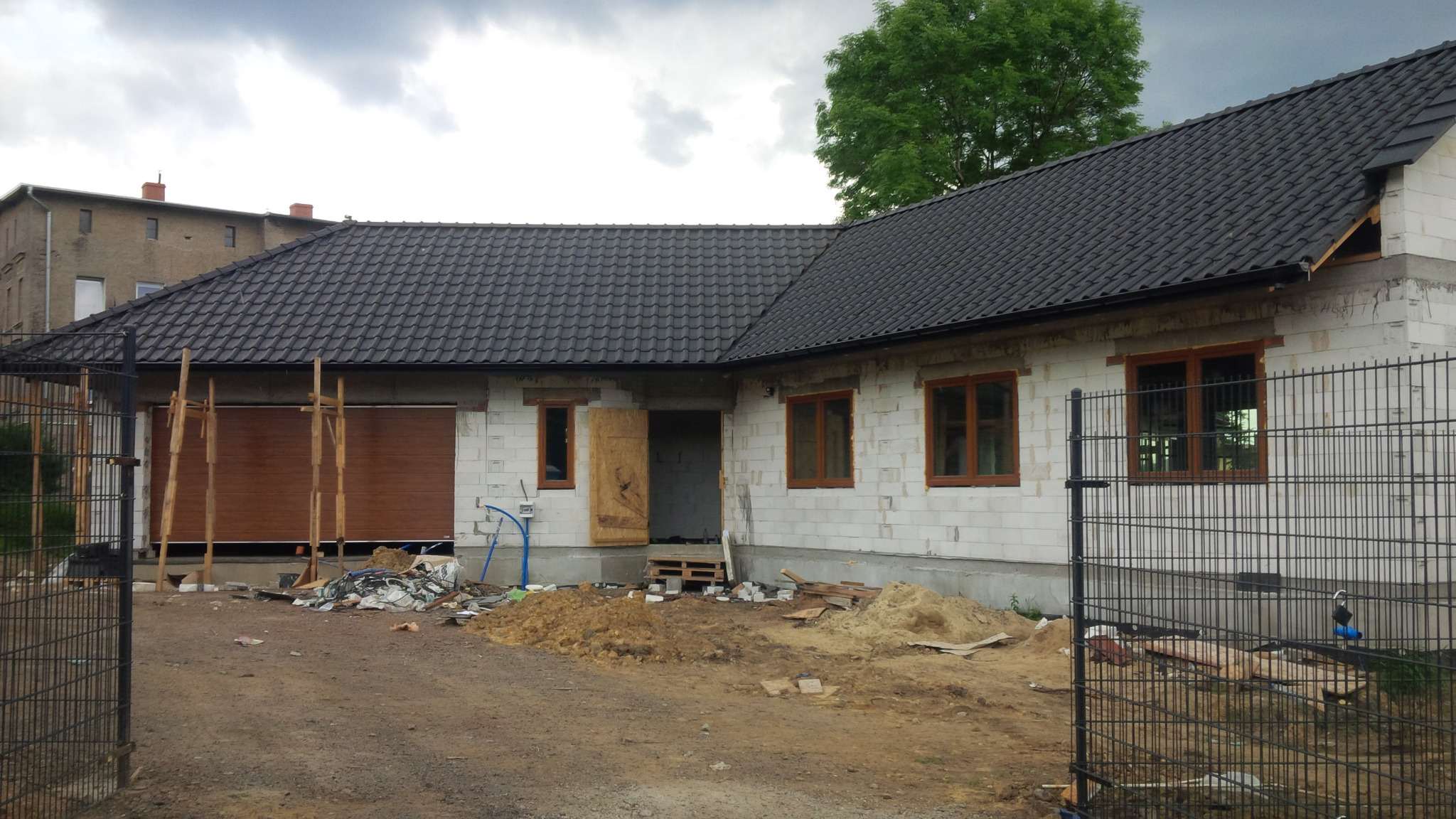 Dom murowany w trakcie budowy: widoczne białe bloczki, ciemna dachówka, drewniane okna i brama garażowa z drewnianą konstrukcją wsporczą. Teren wokół domu częściowo zagospodarowany, widoczny gruz...