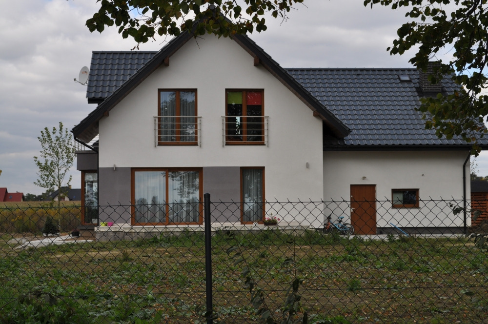 Dwukondygnacyjny dom z białymi ścianami i ciemną dachówką, widoczny zza metalowego ogrodzenia. Na piętrze dwa okna z balkonami, na parterze duże okno tarasowe i drzwi wejściowe. Obok domu...