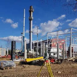 Stanowisko pomiarowe z teodolitem na trójnogu, w tle budowa konstrukcji stalowej z kominami przemysłowymi.