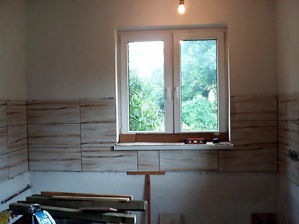 Układanie płytek imitujących drewno na ścianie w pomieszczeniu z oknem, widoczne narzędzia i materiały budowlane.