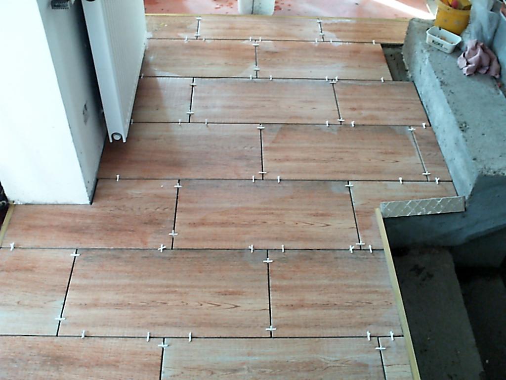 Układanie płytek imitujących drewno na schodach, widoczne krzyżyki dystansowe między płytkami, obok grzejnik i betonowa konstrukcja.