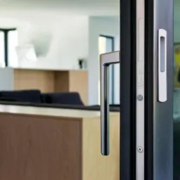 Detal widoku z wnętrza domu na przesuwne okno tarasowe z aluminiową ramą i minimalistyczną, podłużną klamką. W tle widoczny salon z nowoczesnymi meblami.