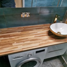 Usługi Remontowo-Budowlane Tip-Top Krzysztof Nidecki - Nowoczesna łazienka z blatem drewnianym nad pralką, umywalką w kolorze złota i baterią. Turkusowe płytki na ścianie i lustro w tle.