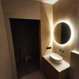 Usługi Remontowo-Budowlane Tip-Top Krzysztof Nidecki - Nowoczesna łazienka z umywalką nablatową, okrągłym lustrem z podświetleniem LED i ścianą w geometryczny wzór. Widoczny fragment toalety i niedokończone wejście.