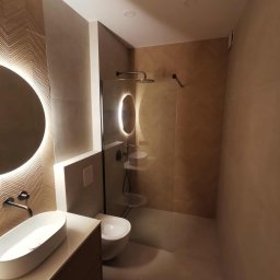 Usługi Remontowo-Budowlane Tip-Top Krzysztof Nidecki - Nowoczesna łazienka z okrągłym lustrem LED, umywalką nablatową i kabiną prysznicową typu walk-in. Ściany wykończone płytkami w odcieniach beżu z subtelnym wzorem jodełki.