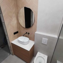 Usługi Remontowo-Budowlane Tip-Top Krzysztof Nidecki - Nowoczesna łazienka z umywalką nablatową, okrągłym lustrem i toaletą podwieszaną. Ściana z wzorem jodełki. Minimalistyczny design, jasne kolory, eleganckie wykończenie.