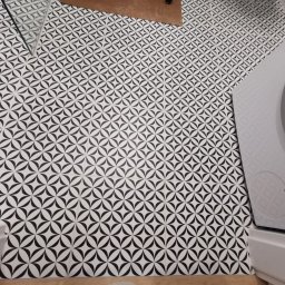 Usługi Remontowo-Budowlane Tip-Top Krzysztof Nidecki - Wzorzyste, czarno-białe płytki podłogowe w łazience, ułożone pod kątem, z widoczną pralką i fragmentem umywalki. Jasne fugi podkreślają geometryczny deseń.