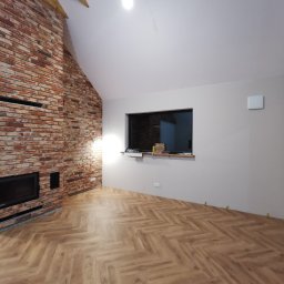 Usługi Remontowo-Budowlane Tip-Top Krzysztof Nidecki - Salon z ceglaną ścianą kominkową i parkietem ułożonym w jodełkę. Widoczne drewniane belki stropowe i okno z widokiem na ceglaną ścianę. Wnętrze w trakcie wykańczania.