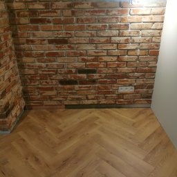 Usługi Remontowo-Budowlane Tip-Top Krzysztof Nidecki - Ukończona ściana z cegły i parkiet ułożony w jodełkę. Widoczne gniazdka elektryczne i oświetlenie punktowe. Detal wykończenia wnętrza.