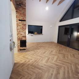 Usługi Remontowo-Budowlane Tip-Top Krzysztof Nidecki - Salon z nową podłogą w jodełkę, ceglanym kominem i surowym drewnem na suficie. Widoczne okno i drzwi balkonowe. Kabel wystaje ze ściany.