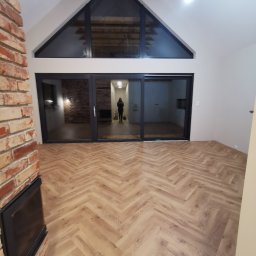 Usługi Remontowo-Budowlane Tip-Top Krzysztof Nidecki - Jasne panele ułożone w jodełkę francuską w przestronnym salonie z ceglanym kominkiem i dużym oknem. Widoczne drewniane belki stropowe i przesuwne drzwi tarasowe.