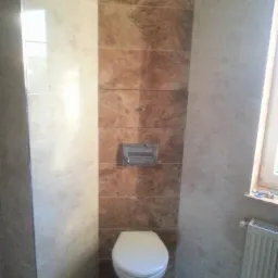 Wykończona łazienka z toaletą, ściany wyłożone płytkami w odcieniach beżu i brązu imitującymi marmur, widoczny grzejnik pod oknem.