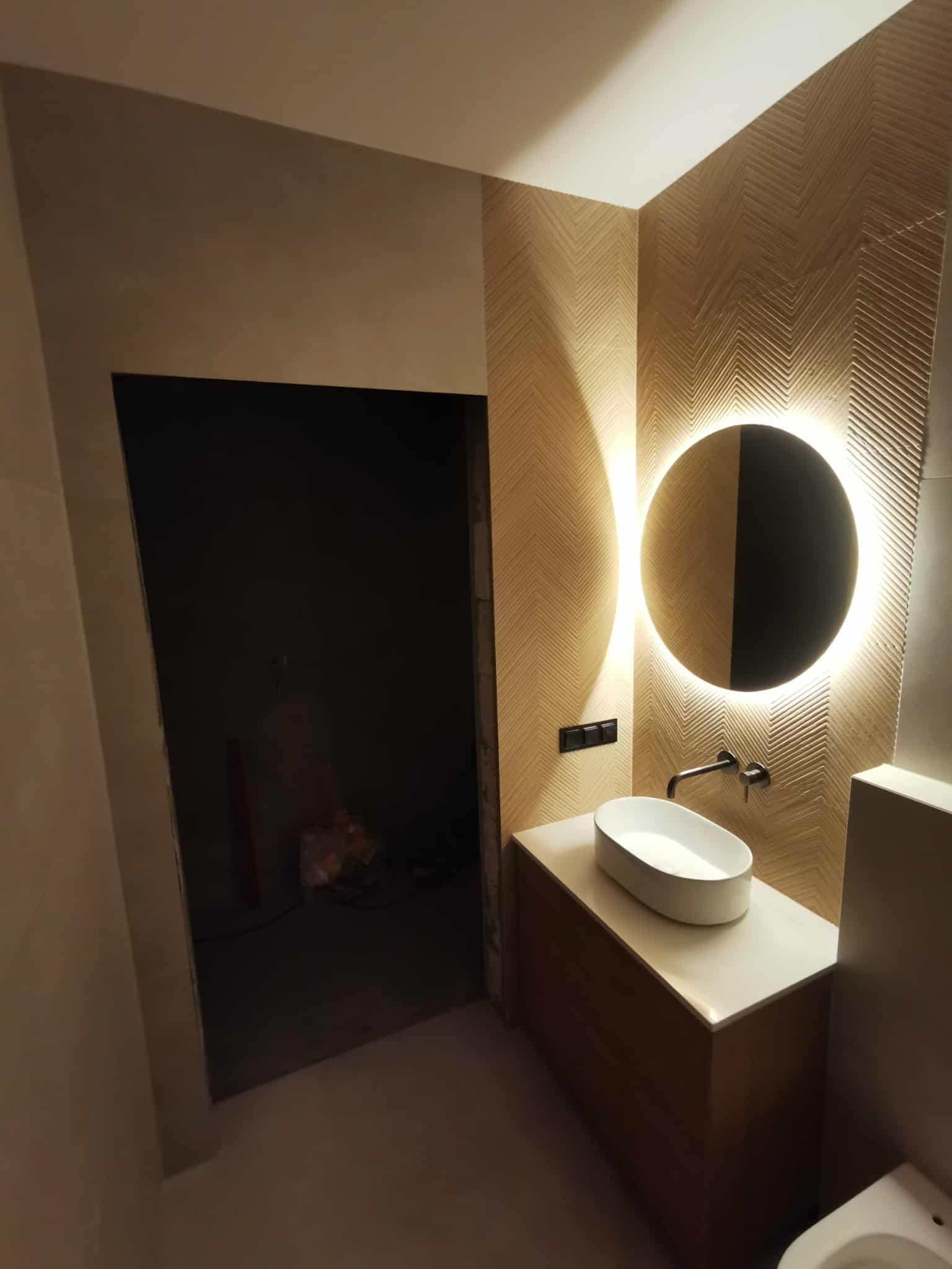 Nowoczesna łazienka z umywalką nablatową, okrągłym lustrem z podświetleniem LED i ścianą w geometryczny wzór. Widoczny fragment toalety i niedokończone wejście.