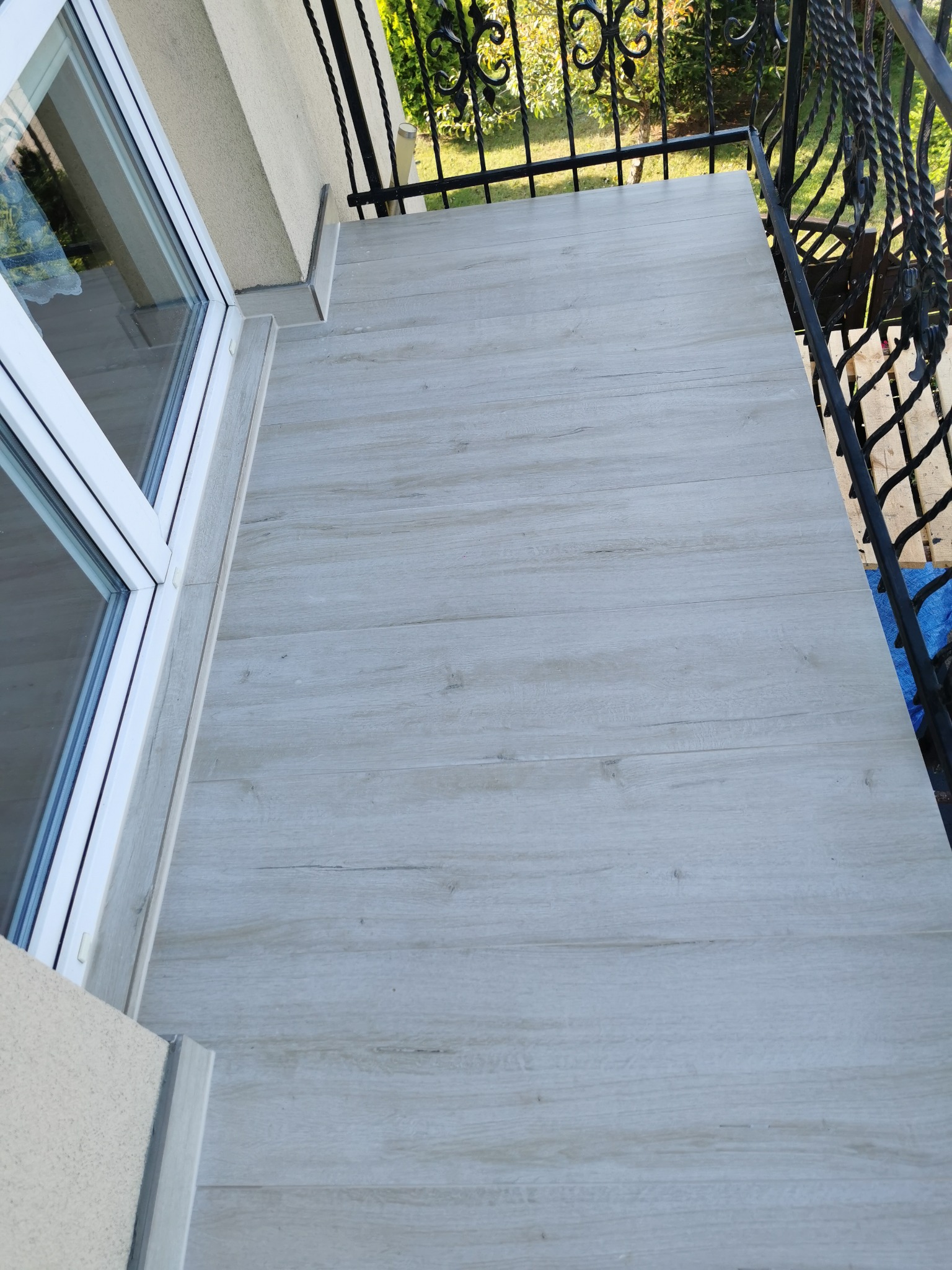 Jasnoszara podłoga z paneli na balkonie z czarną, kutą balustradą. Widok z góry, ukośny kadr. Nowoczesne wykończenie balkonu z drewnopodobnymi panelami.