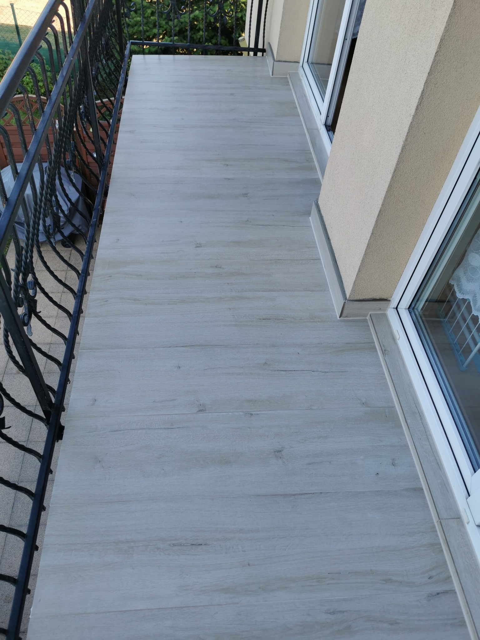 Balkon z podłogą wyłożoną jasnymi panelami imitującymi drewno, z czarną, ozdobną balustradą i widocznym fragmentem okna. Ujęcie z góry, podkreślające wzór podłogi.