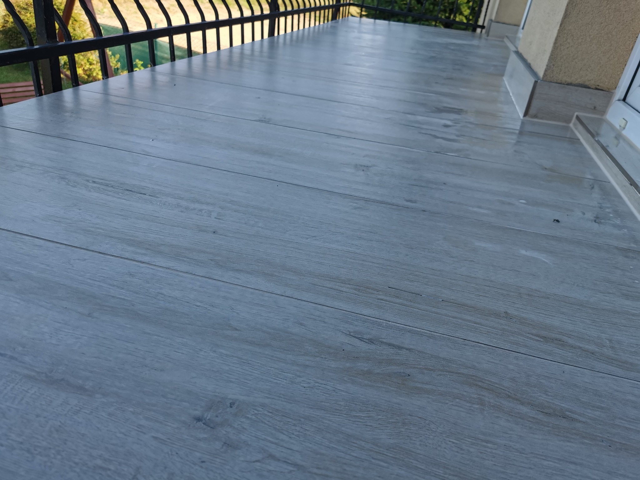 Jasnoszara podłoga z desek na balkonie z czarną balustradą. Widok z góry, ukośny kadr. Naturalne światło podkreśla strukturę drewna. Detal wykończenia.