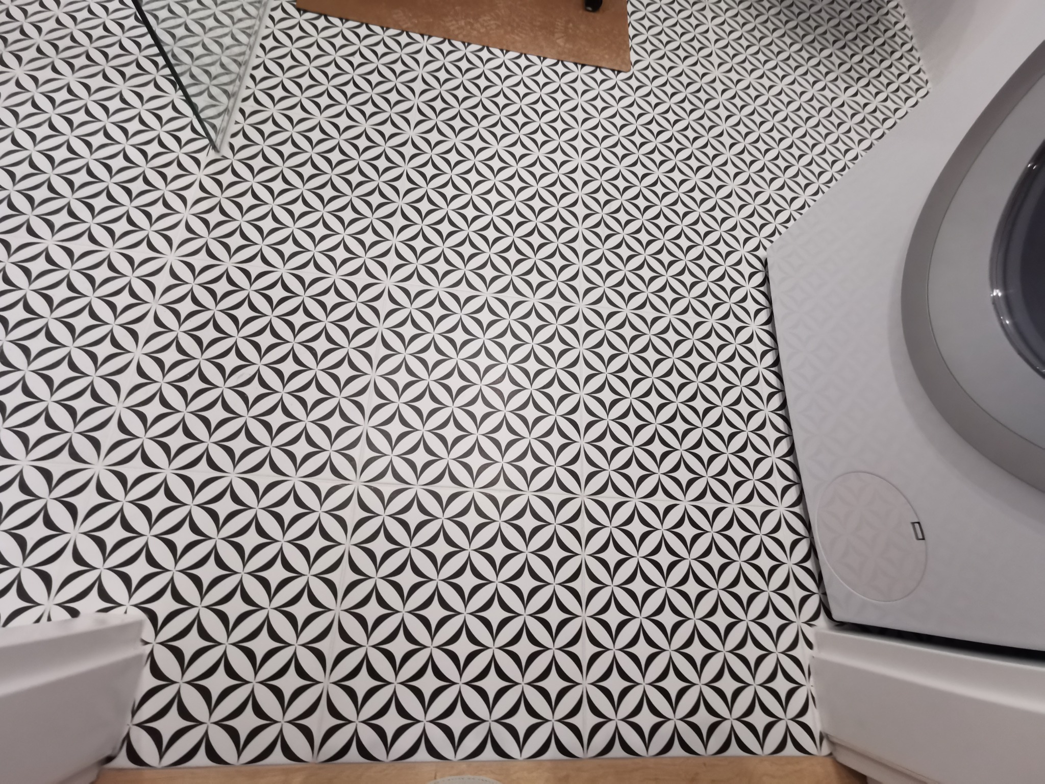 Wzorzyste, czarno-białe płytki podłogowe w łazience, ułożone pod kątem, z widoczną pralką i fragmentem umywalki. Jasne fugi podkreślają geometryczny deseń.