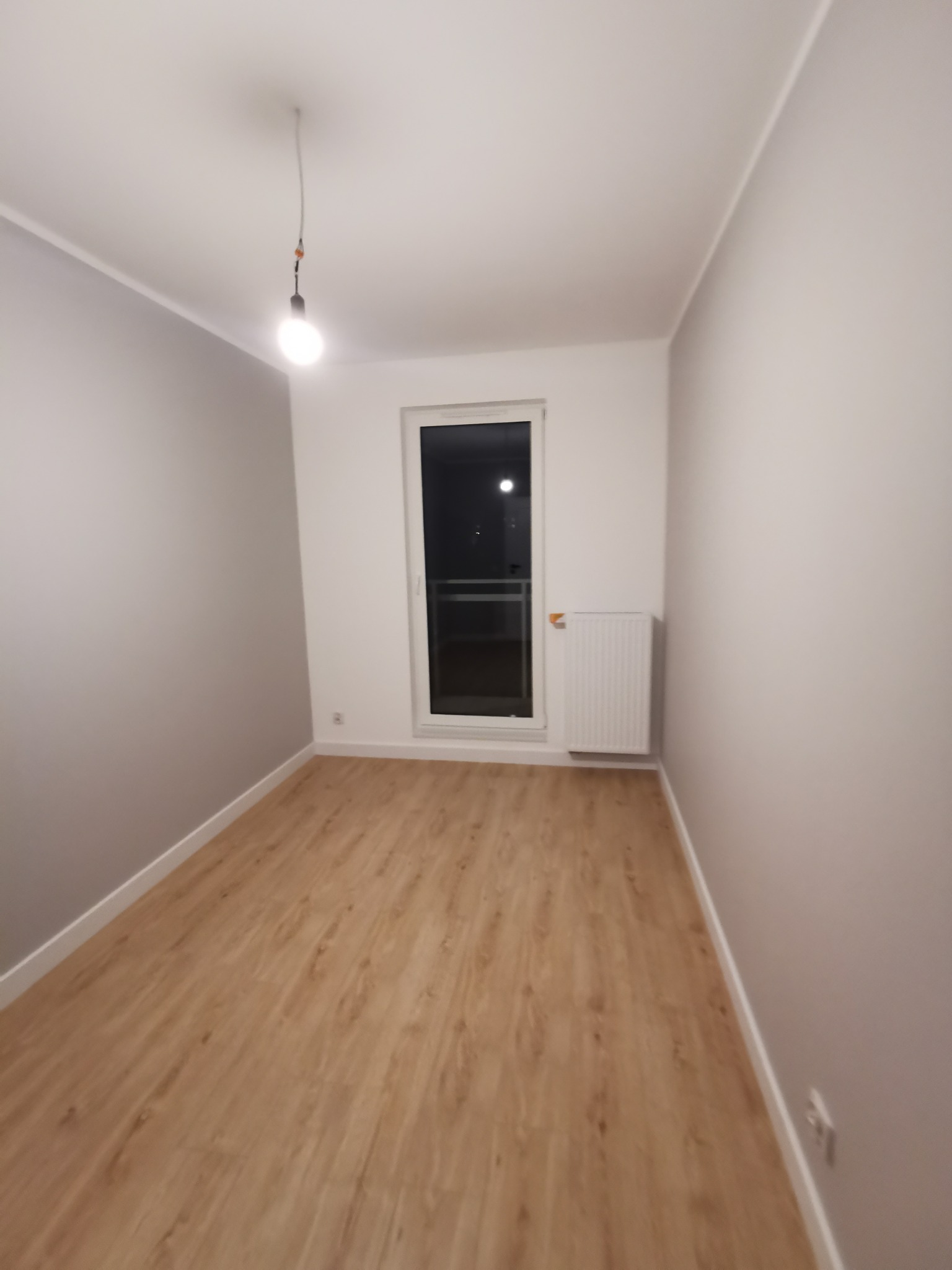 Pomalowane na szaro ściany i ułożone panele podłogowe w pustym pokoju z oknem i grzejnikiem. Wisząca żarówka. Wykończenie wnętrza w nowym budownictwie.