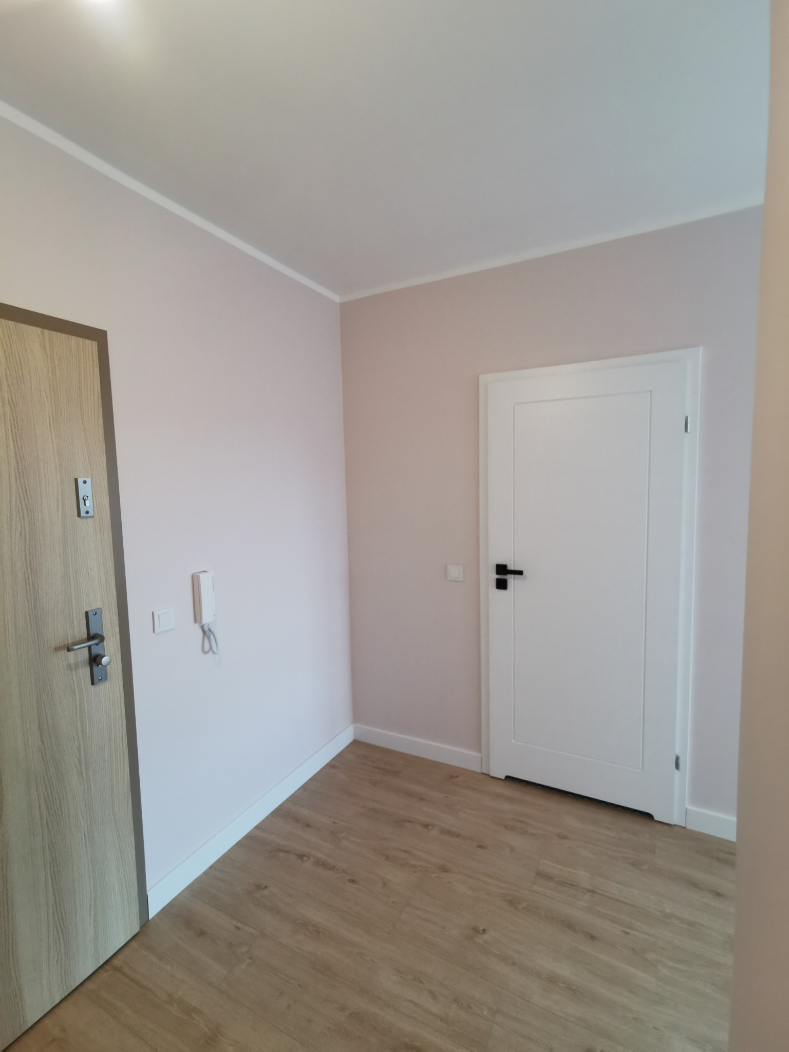 Jasne wnętrze po remoncie: drewnopodobna podłoga, ściany w odcieniach różu, białe drzwi wewnętrzne i wejściowe z elementami drewna. Minimalistyczny design.