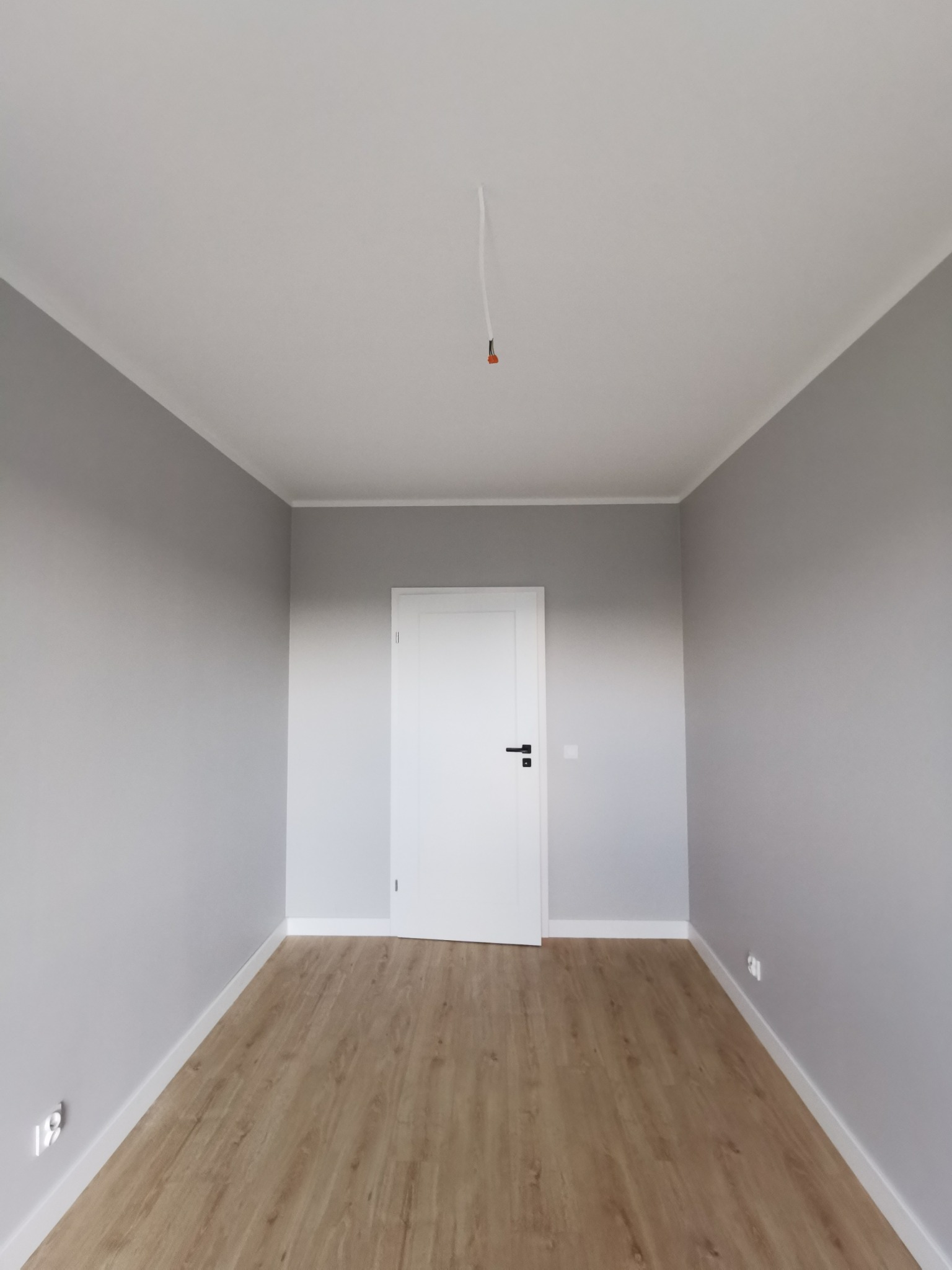 Pokój po remoncie: szare ściany, drewnopodobna podłoga, białe listwy przypodłogowe i drzwi. Widoczny wystający kabel z sufitu. Minimalistyczny design.