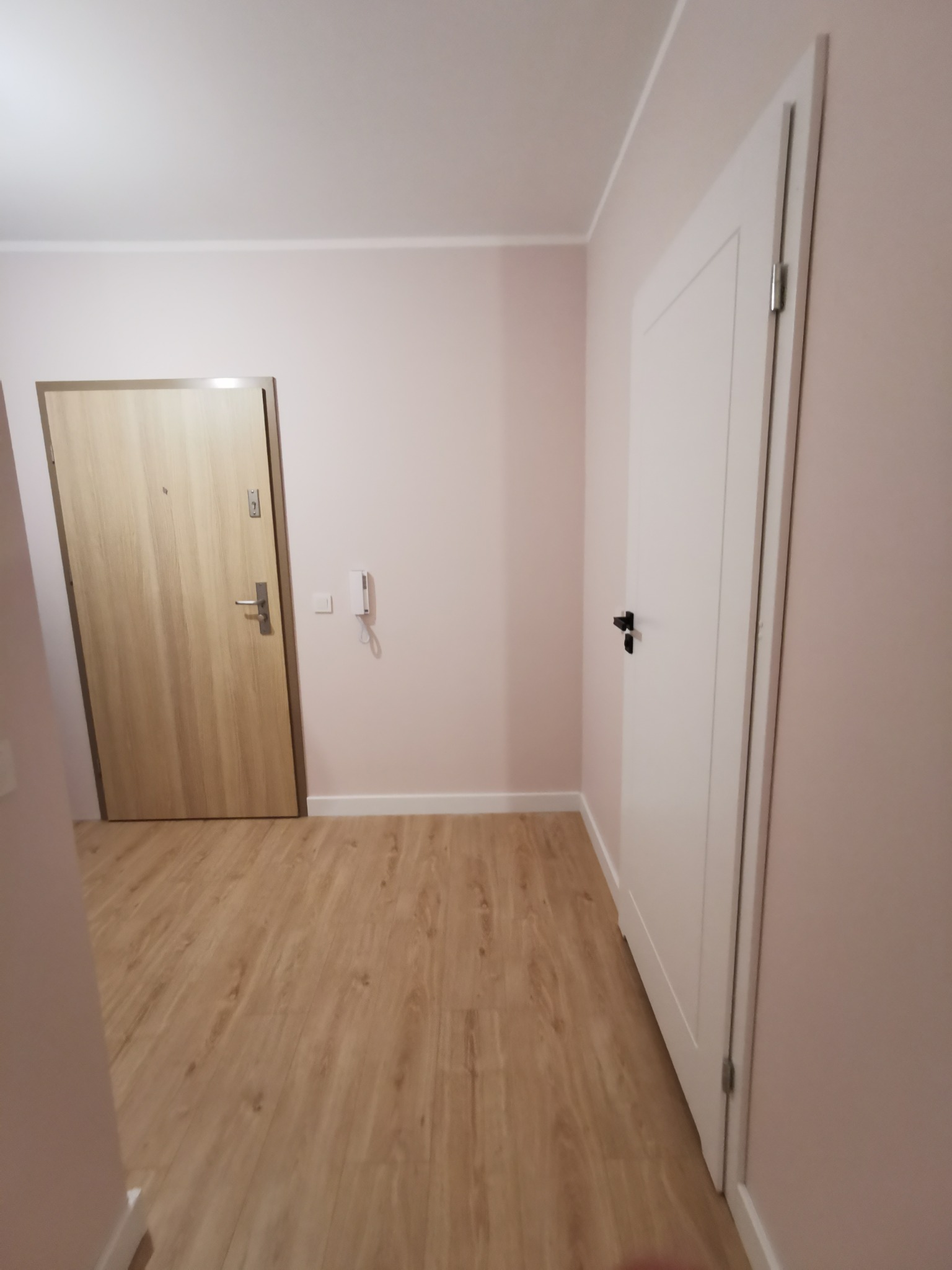 Jasne panele podłogowe w korytarzu z dwojgiem drzwi: wejściowymi w odcieniu drewna i wewnętrznymi, białymi z czarną klamką. Ściany w delikatnym, pudrowym różu.