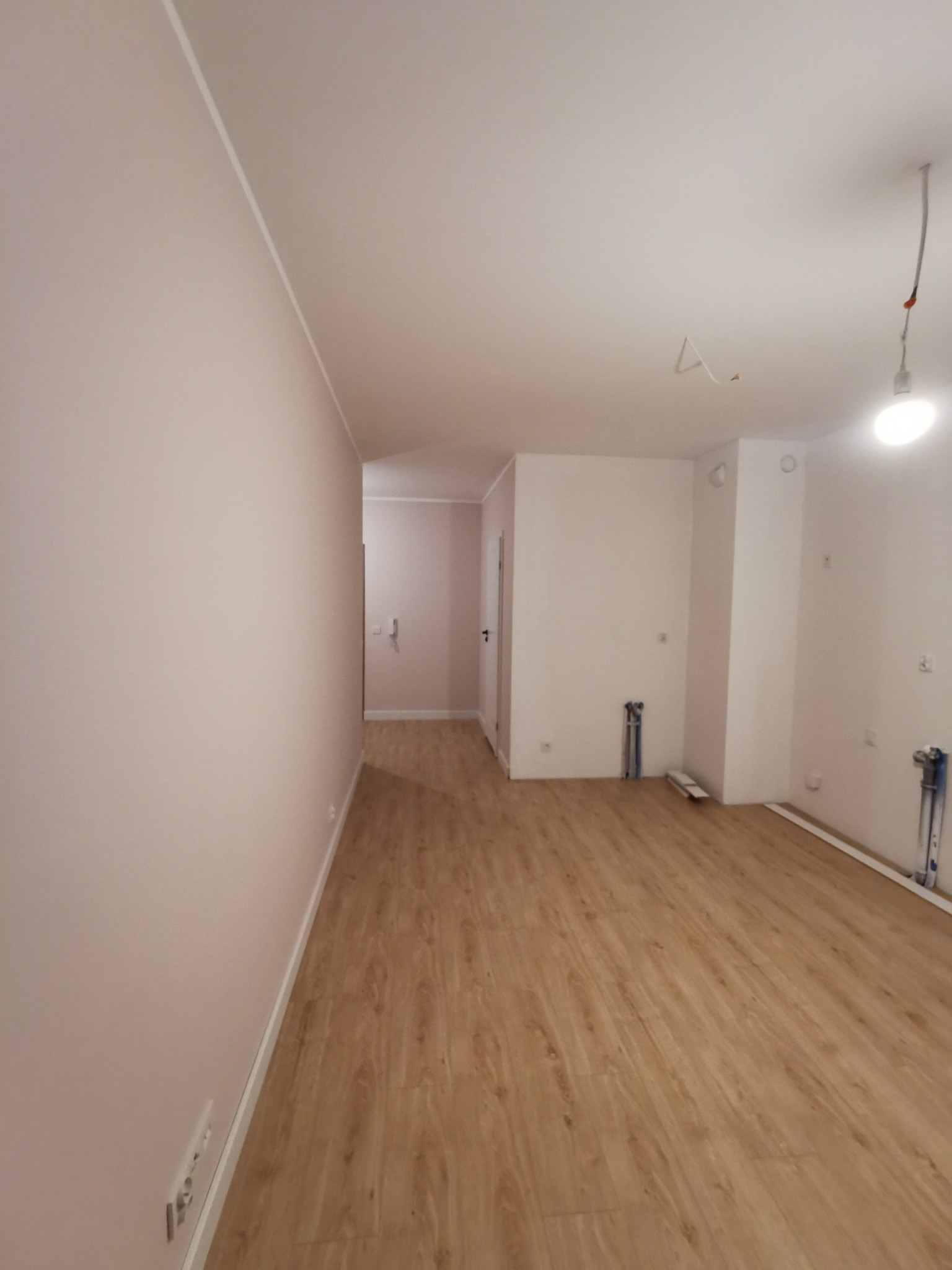 Jasne panele podłogowe w nowym wnętrzu po remoncie. Ściany w odcieniach bieli i delikatnego różu. Widoczne instalacje wodne i elektryczne przed wykończeniem.
